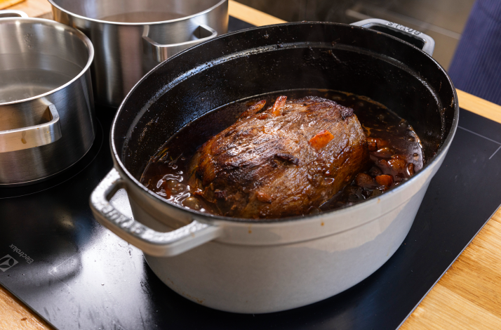 Beef roast simmering in rich pašticada sauce within a white cast-iron pot on a kitchen stovetop.