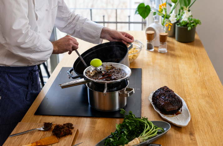 Chef stirring rich pašticada braising sauce over induction hob, with seared beef joint and fresh herbs nearby.