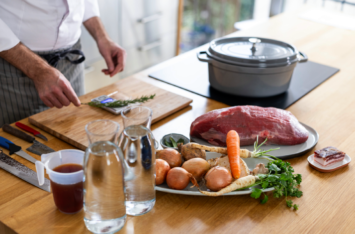 Chef prepping pašticada ingredients: raw beef, onions, carrot, parsley root, herbs, red wine, and water on a counter.