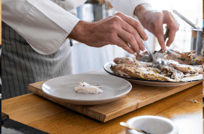 Chef plating Dalmatian fish soup, portioning poached fish onto a grey plate from a serving platter.