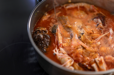 Steam rising from a simmering brudet stew in a steel pot, with prawns, fish, and squid in tomato-based broth.