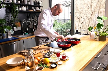 Chef in white coat preparing Dalmatian marinated fish in red pan, with fresh ingredients on wooden kitchen counter.