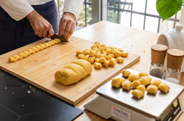 Chef slicing gnocchi dough rope into portions on a wooden board, with shaped pieces and a kitchen scale alongside.