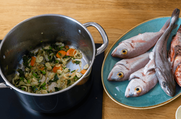 A large saucepan with sauté vegetables and a plate next to it with fresh fish – ingredients for Dalmatian fish soup.