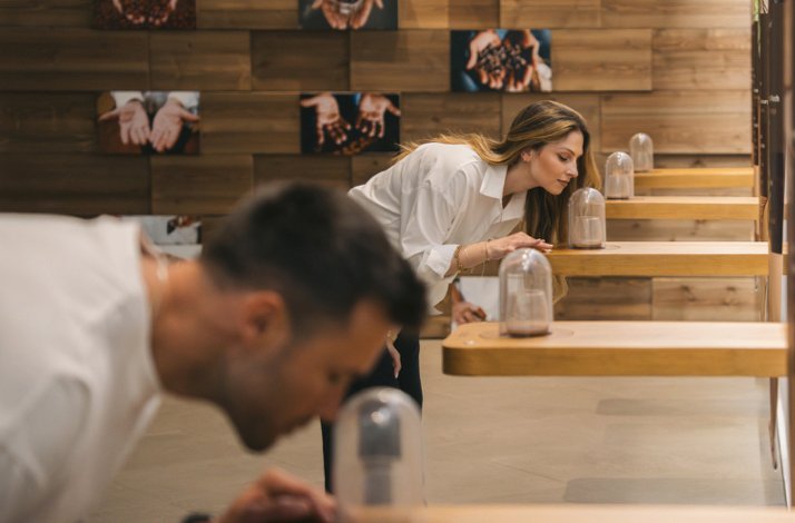 Two visitors leaning over tiered wooden shelves to smell glass-domed aroma samples in a sensory exhibition room at The Wine Experience - Wine Museum.