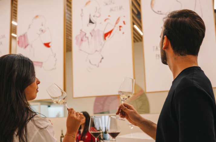 Two visitors smelling white wine during a guided tasting session at The Wine Experience - Wine Museum, with illustrated artworks on the gallery walls.