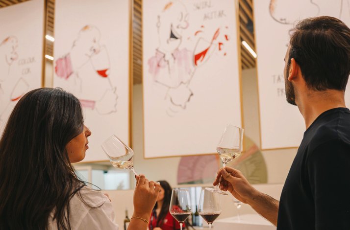 Two visitors smelling white wine during a guided tasting session at The Wine Experience - Wine Museum, with illustrated artworks on the gallery walls.