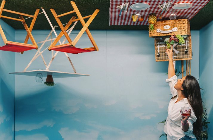 Visitor reaching toward an inverted picnic installation inside a The Wine Experience - Wine Museum gallery, with suspended basket and chairs overhead.