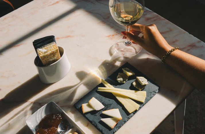 Hand lifting a white wine glass beside a curated cheese and cracker board served on a marble table at The Wine Experience - Wine Museum.