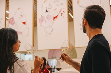 Two visitors smelling white wine during a guided tasting session at The Wine Experience - Wine Museum, with illustrated artworks on the gallery walls.