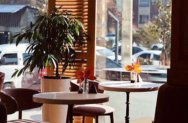 Window-side seating area at Teplo Cafe, Tashkent, with round marble tables, potted plant, and street view through large windows.