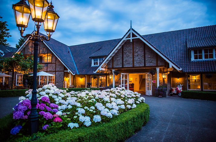 Exterior of Landhuishotel De Bloemenbeek in the Netherlands at dusk with glowing street lamps and blooming hydrangeas.