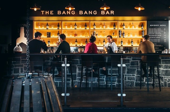 Bartender serving guests at The Bang Bang Bar inside Teeling Whiskey Distillery, with a backlit whiskey bottle display.