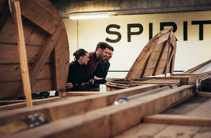 Three distillery workers peering into open wooden fermentation vessels, with 'SPIRIT' painted on the wall behind them.