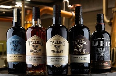 Five bottles of Teeling Irish Whiskey variants standing on a wooden surface against a distillery background.