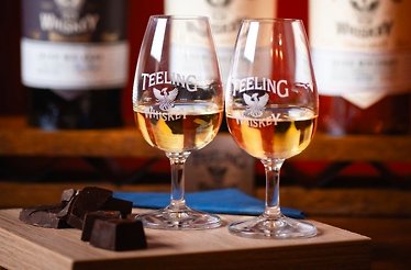 Two branded Teeling whiskey glasses and dark chocolate on a wooden board, with bottles in the background.