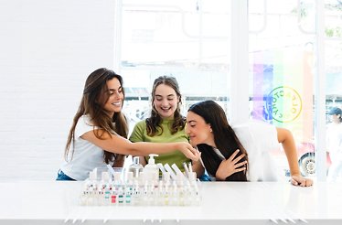 Three smiling women leaning together over a white counter of fragrance vials at Olfactory NYC, one holding a scent strip toward another to smell.