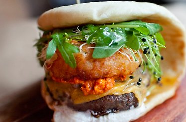 Bäco sandwich from le dräQ, layered with beef patty, red harissa, melted cheese, crispy onion, microgreens, and pea shoots.