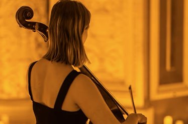 A woman from behind playing a cello in a golden, candlelit room.
