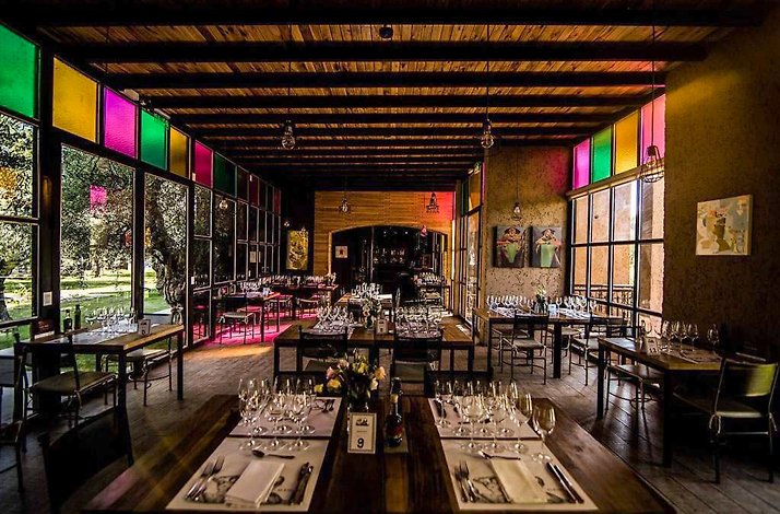 Casa Vigil - Interior dining room with a wood-beamed ceiling, colored stained-glass panels above large windows, and long communal wooden tables set with wine glasses, place settings and floral centerpieces; garden visible outside.