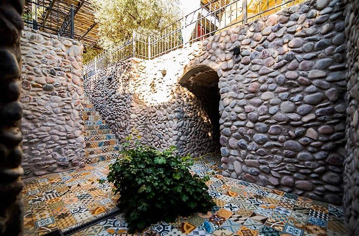 Casa Vigil - Outdoor stone courtyard with rounded river-stone walls, an arched passage and a tiled staircase, floored in colorful patterned encaustic cement tiles, with a small green shrub at the center.