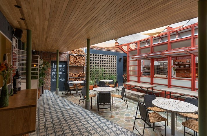 Interior courtyard dining room at Mesa Franca, Bogotá, Colombia — stacked firewood, a red steel-framed open kitchen, terrazzo tables with woven bistro chairs and patterned tile flooring under a timber ceiling
