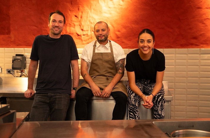 The Mesa Franca team at the kitchen pass in Bogotá, Colombia — an aproned chef flanked by two colleagues against a terracotta wall above white metro-tiled walls and a stainless-steel kitchen counter