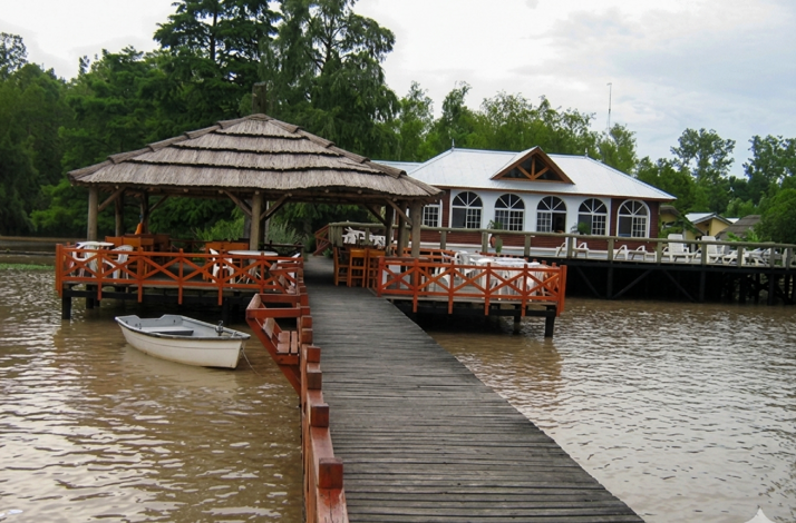 Wooden dock leading to a thatched-roof gazebo and a riverside lodge, with a small rowboat moored at the edge.