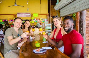 Food tour guide explaining local flavors to a small group seated outdoors in Playa del Carmen, surrounded by leafy trees and colorful street setting.