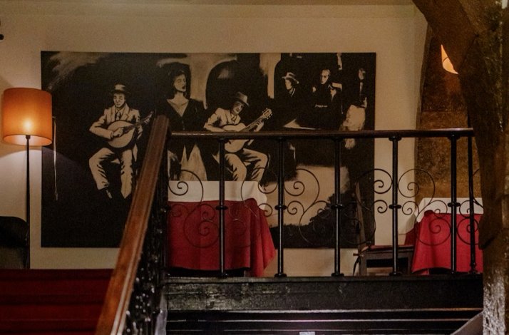 Black-and-white fado mural above a wrought-iron staircase railing at Clube de Fado, Lisbon, with red-draped tables below.