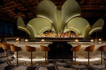 Grand curved bar at Lucia LA topped by an illuminated petal-shaped installation above a full spirits display.