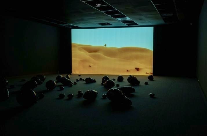 A projection of a desert landscape is displayed in a dark exhibition hall. In the foreground, dark gourd-like objects are scattered on the floor