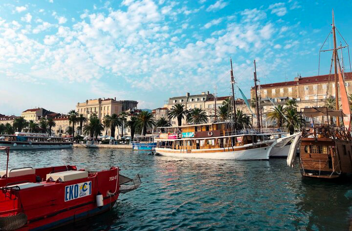 Boats and yachts docked along the palm-lined Riva promenade under a partly cloudy sky in Split.