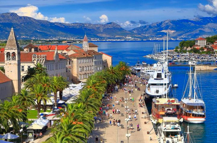 High-angle view of Trogir's historic waterfront, featuring a bell tower, palm trees, and yachts.