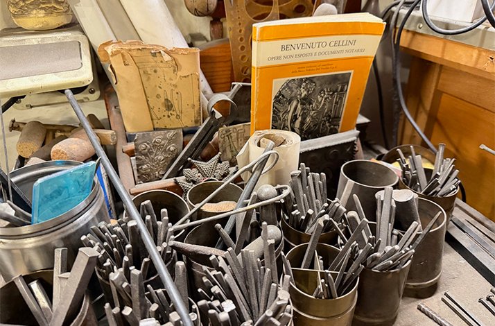 A collection of traditional crafting tools and materials in an artisan's workshop, with a book on Benvenuto Cellini displayed in the background.