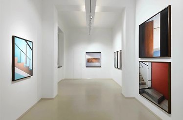 Interior of a contemporary art gallery in Rome, featuring framed artworks on white walls and a large window overlooking lush greenery.