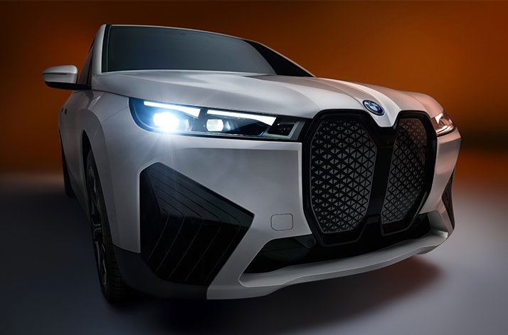 Silver BMW SUV front view with headlights on against a warm brown background.