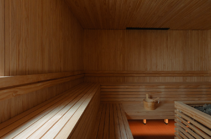 Empty wood-panelled sauna room with tiered benches, a small wooden bucket on a stool, and sauna stones beside a heater.
