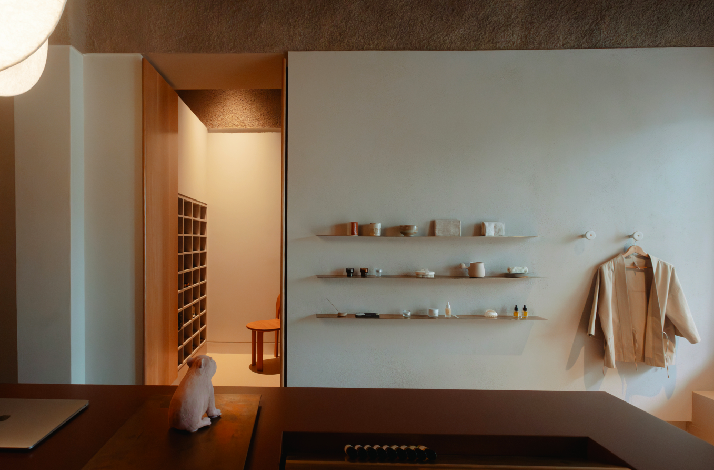 Minimalist spa lounge displaying ceramic wares on floating wall shelves, a hanging robe, and a white capybara figurine on a wooden desk.