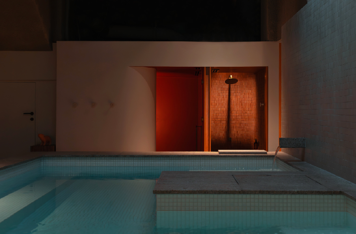Dimly lit indoor pool area featuring a warmly glowing shower alcove with terracotta tiles and overhead rainfall showerhead.