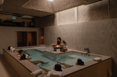 Wellness practitioner playing a handpan over a warm soaking pool while robed guests relax along the pool surround indoors.