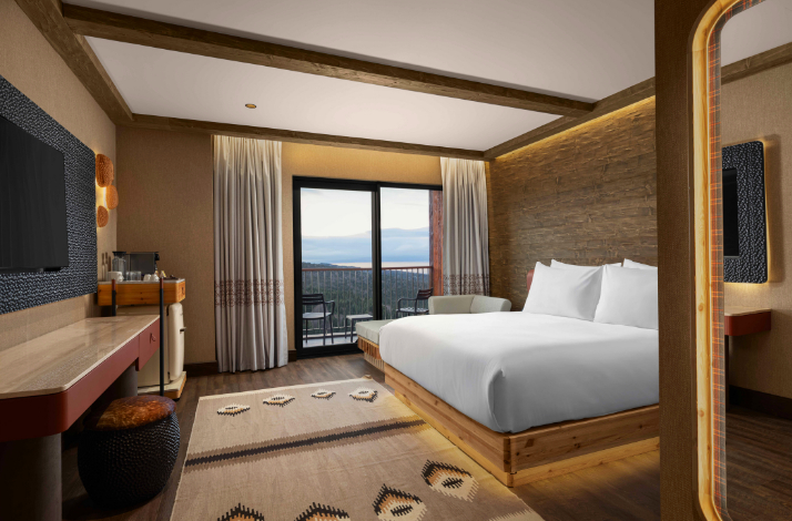 Modern hotel room with a king-size bed, wooden accents, a balcony overlooking a forest, a seating area, a work desk, a TV, and a coffee station.