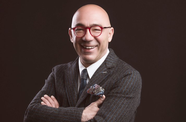 A smiling man wearing glasses and a dark suit, posing with his arms crossed against a dark background.