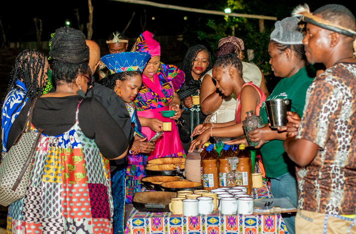  Group of women in colorful traditional South African attire gathered around a table sampling local drinks and foods at night.