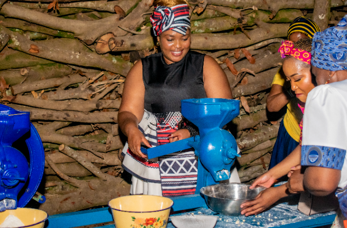 South African woman in traditional Xhosa attire operating a blue hand grain mill during a cultural village experience.
