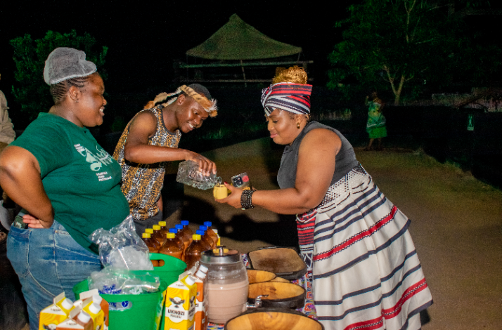 Women in South African traditional and casual attire serving drinks and food at an outdoor cultural evening event.
