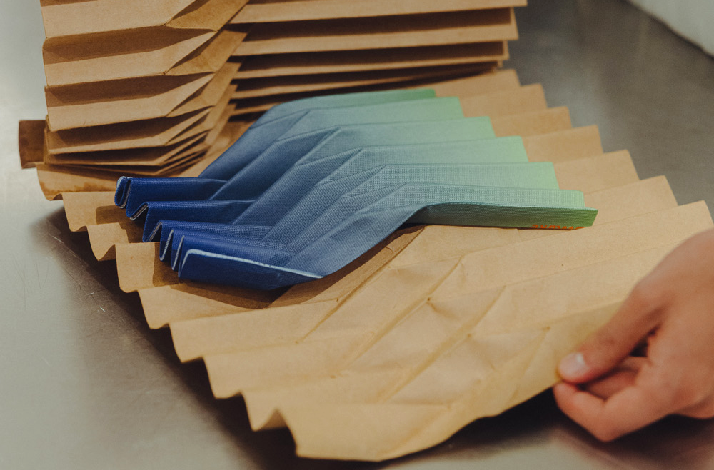 A close-up on a table showing brown paper bags fanned out in neat stacks while a hand adjusts one bag; folded blue-to-green cloth pieces rest on the bags.