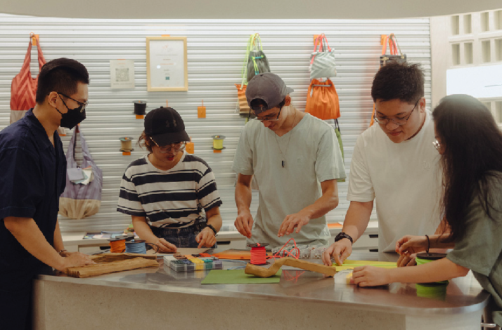Five people collaborate around a workshop table, working with colorful cords and materials; tote bags hang on the wall behind them.