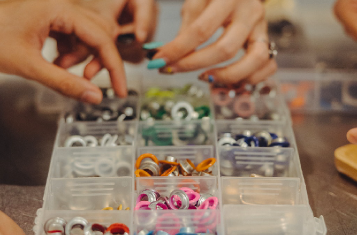 Hands sort colorful metal grommets and rings in a clear plastic compartment organizer.