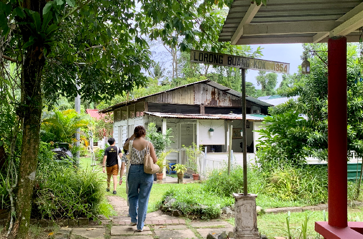 Visitors strolling through Kampong Lorong Buangkok's lush village lane, passing the historic 'Lorong Buang Kok 1954' street sign amid traditional wooden houses.